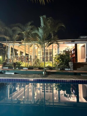 A nighttime view of a hotel with a swimming pool, palm trees, and a well-lit entrance. - Kindo Kicha Farmhouse (Moshi)