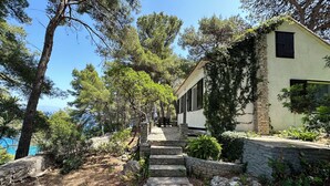 Un chemin de pierres mène à un bâtiment blanc aux volets noirs, entouré d’arbres et d’un mur de pierres.