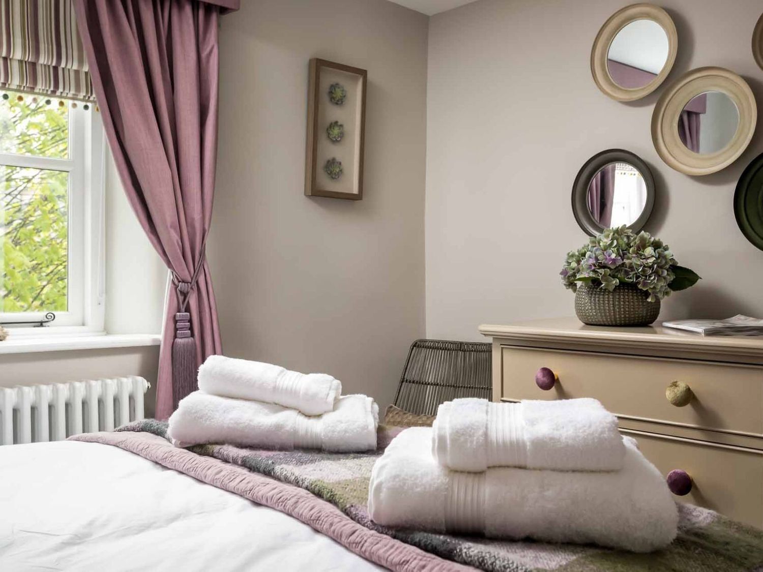 A neatly made bed with folded towels, a radiator, a window with curtains, and a dresser with a plant and mirrors.