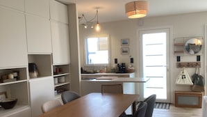 A modern kitchen with white cabinetry, a wooden dining table, and chairs. There is a window providing natural light, and a kitchen island with a sink and countertop.