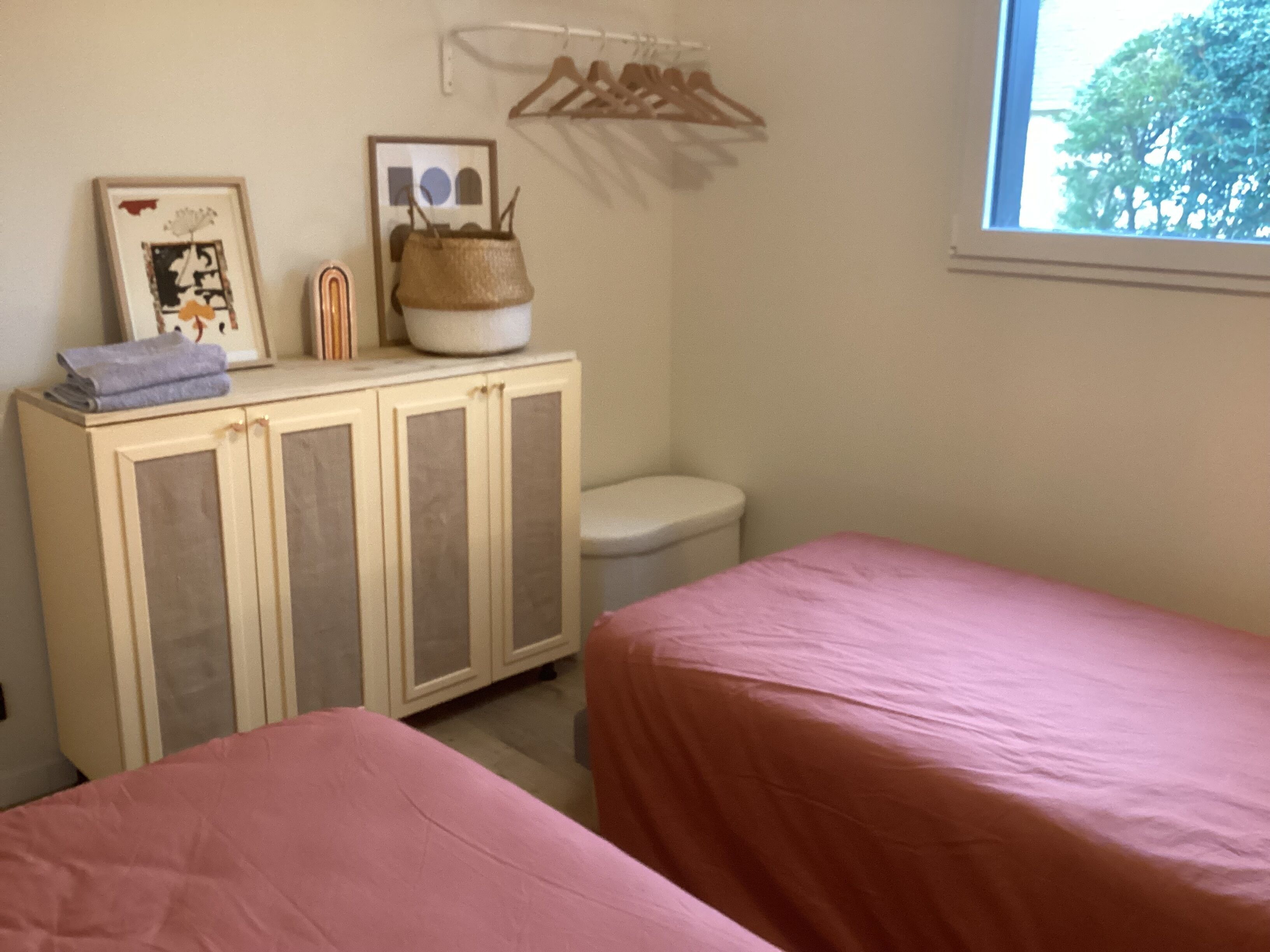 A bedroom with two beds, a small cabinet, a hanging clothes rack, and a window with a view of trees.
