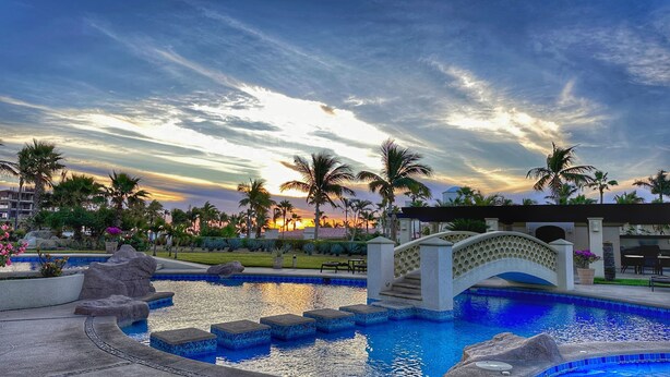 A resort pool area with a bridge, palm trees, and a sunset view. - Las Mananitas Luxury Triple en-suite Ocean & Beach Front Resort Condo (San José del Cabo)