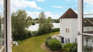 A view of a white building with a brown roof, surrounded by greenery and a body of water.