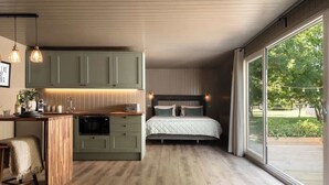 A modern bedroom with a large window, a wooden floor, a bed, and a kitchen area with green cabinets and pendant lighting. - Rabbits Warren - Cotswolds Stay with Sauna and Hot Tub (Wiltshire)