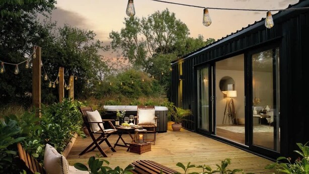 A modern outdoor patio with a wooden deck, seating area, and a glass-enclosed room. - Rabbits Warren - Cotswolds Stay with Sauna and Hot Tub (Wiltshire)