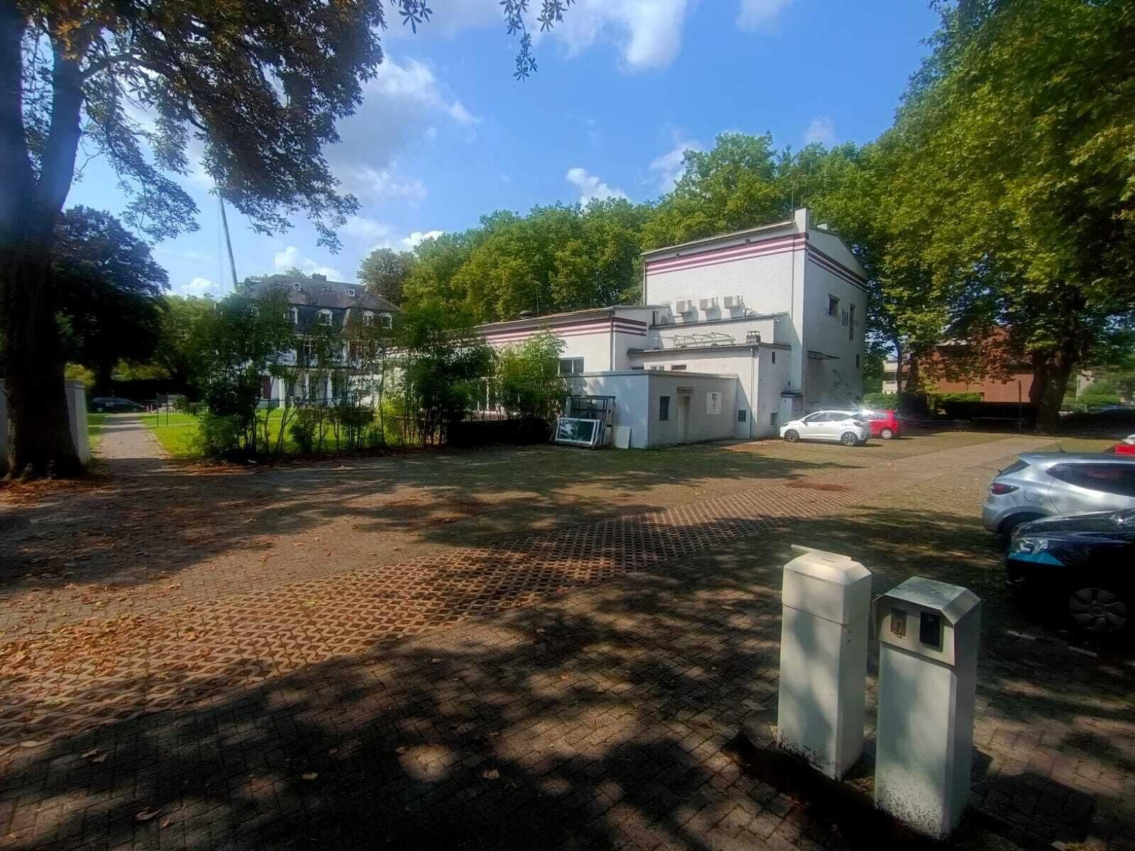 Un edificio con techo plano y fachada blanca, rodeado de árboles y coches estacionados.