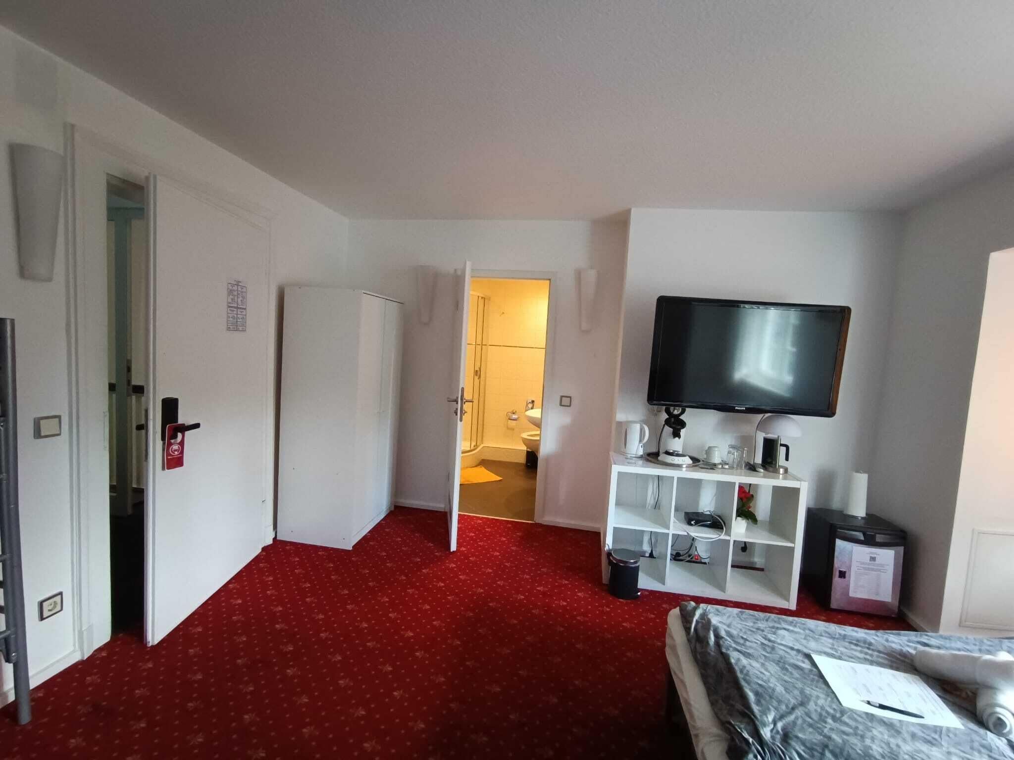 A hotel room with a red carpet, a bed, a TV on a stand, a white cabinet with shelves, and a door leading to a bathroom.