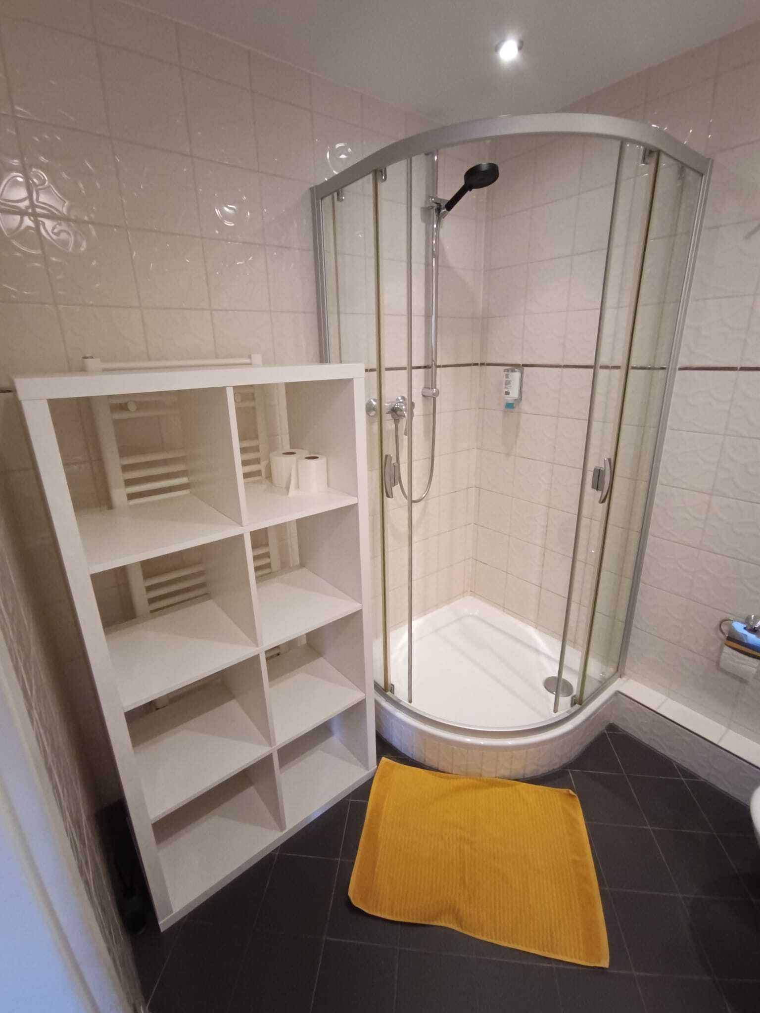 Eine Eckdusche mit Glastür, ein weißes Ablageelement mit Regalen und eine gelbe Badmatte auf dem Boden.
