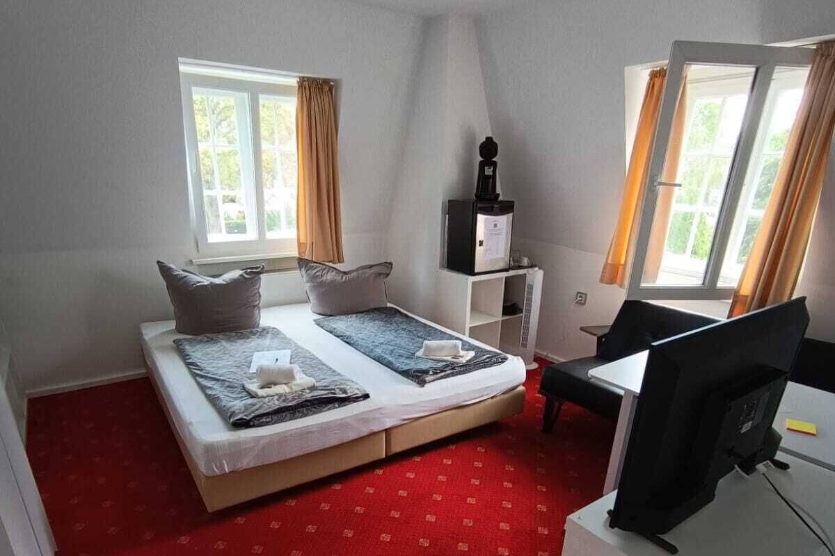 Ein Zimmer mit einem Bett, einem Schreibtisch und einem Computer. Es gibt Fenster mit Vorhängen.