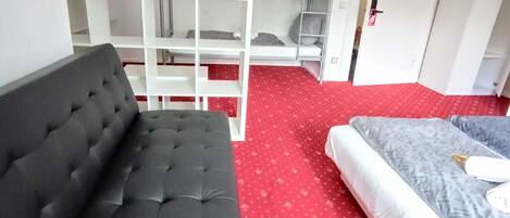 Una habitación con un sofá negro, una cama con colchón gris y un estante blanco.