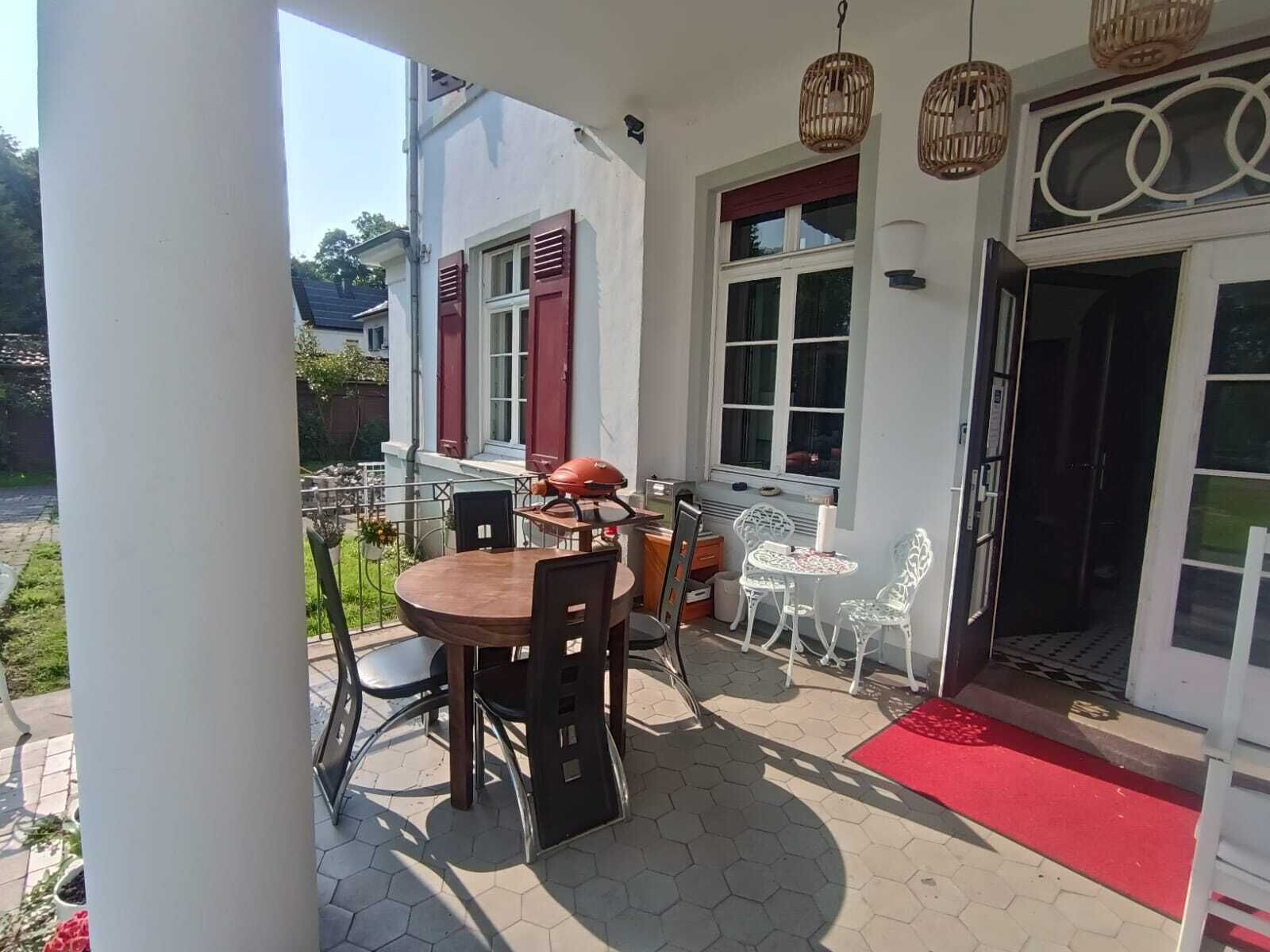 Een overdekt terras met een ronde houten tafel en stoelen, een barbecue en een kleine witte tafel met stoelen.