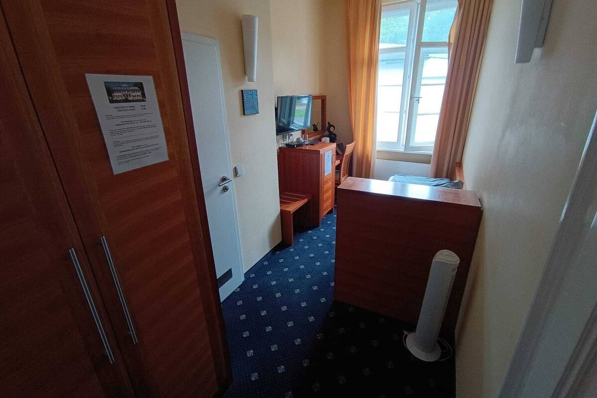 Ein Hotelzimmer mit Holztür, Schreibtisch inklusive Computer, kleinem Fernseher und einem Fenster mit Vorhängen.