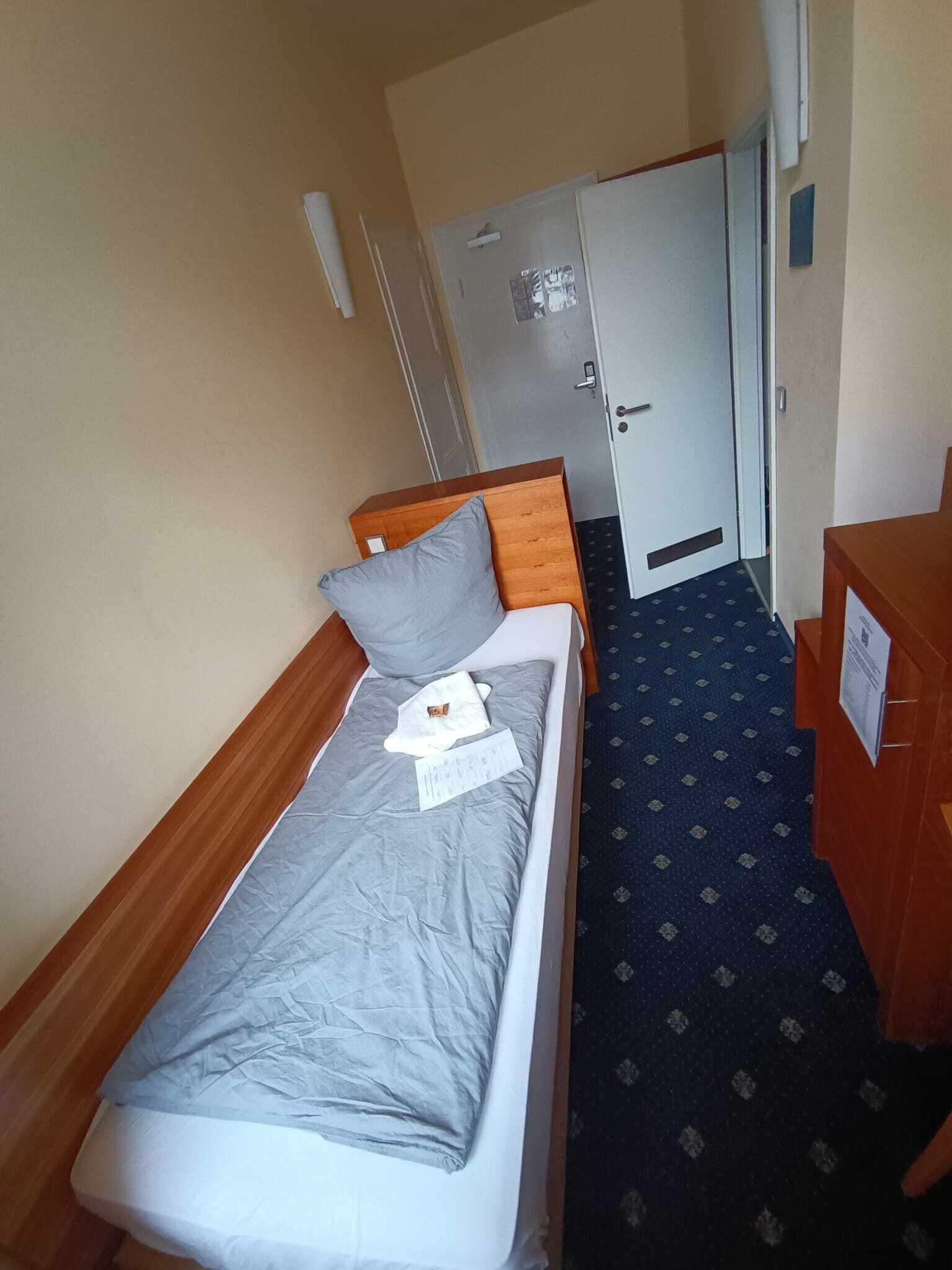 A small, neatly made bed with a grey sheet and pillow, a wooden headboard, and a small wooden dresser to the right.