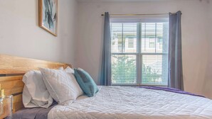 Una cama bien hecha con un cabecero de madera, una mesita de noche con un recipiente de vidrio y una ventana con cortinas azules.