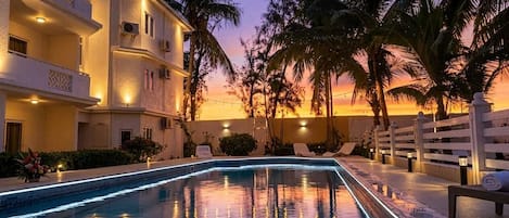 A swimming pool with a night-time lighting feature, surrounded by palm trees and a building with balconies.