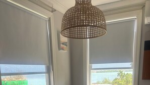 A dining area with a round table, chairs, a woven pendant light, and two windows with a view of the sea and greenery.