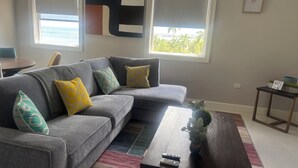 A modern living room with a grey sectional sofa, colorful cushions, a wooden coffee table, and a view of the outdoors.