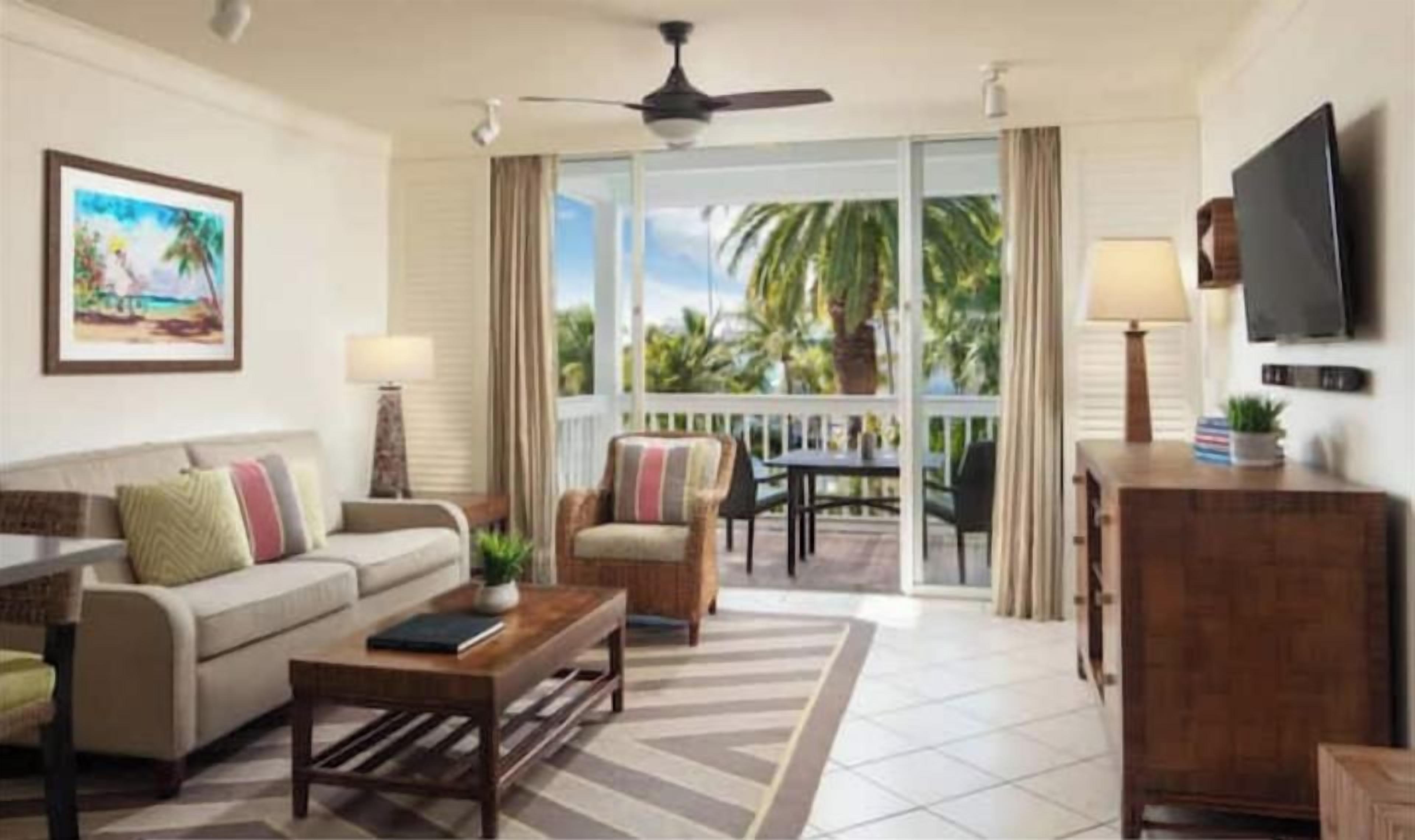 A living room with a sofa, armchairs, a coffee table, and a TV. There's a balcony with a view of palm trees.