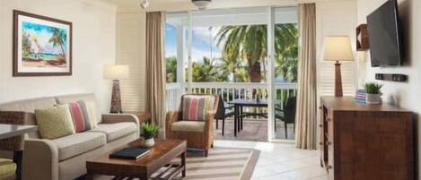 A living room with a sofa, armchairs, a coffee table, and a TV. There's a balcony with a view of palm trees.