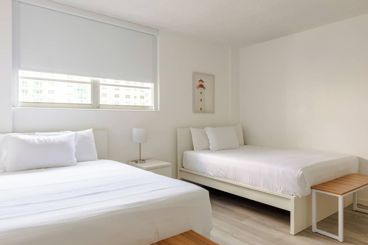 Un dormitorio con dos camas, una mesita de noche con lámpara, un pequeño banco de madera y un cuadro enmarcado en la pared.