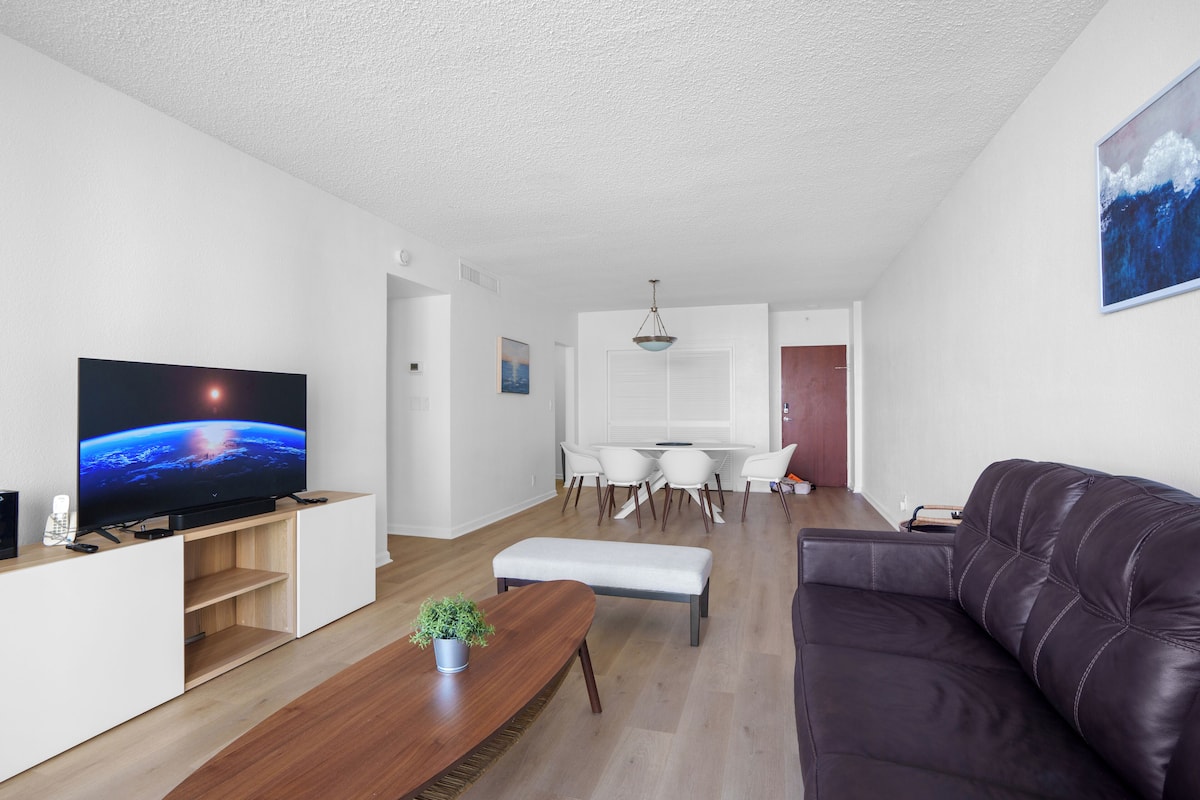 Una sala de estar moderna con un televisor de pantalla plana, mueble de entretenimiento de madera, un sofá de cuero oscuro, una zona de comedor con una mesa y sillas blancas, y una pequeña planta en maceta.