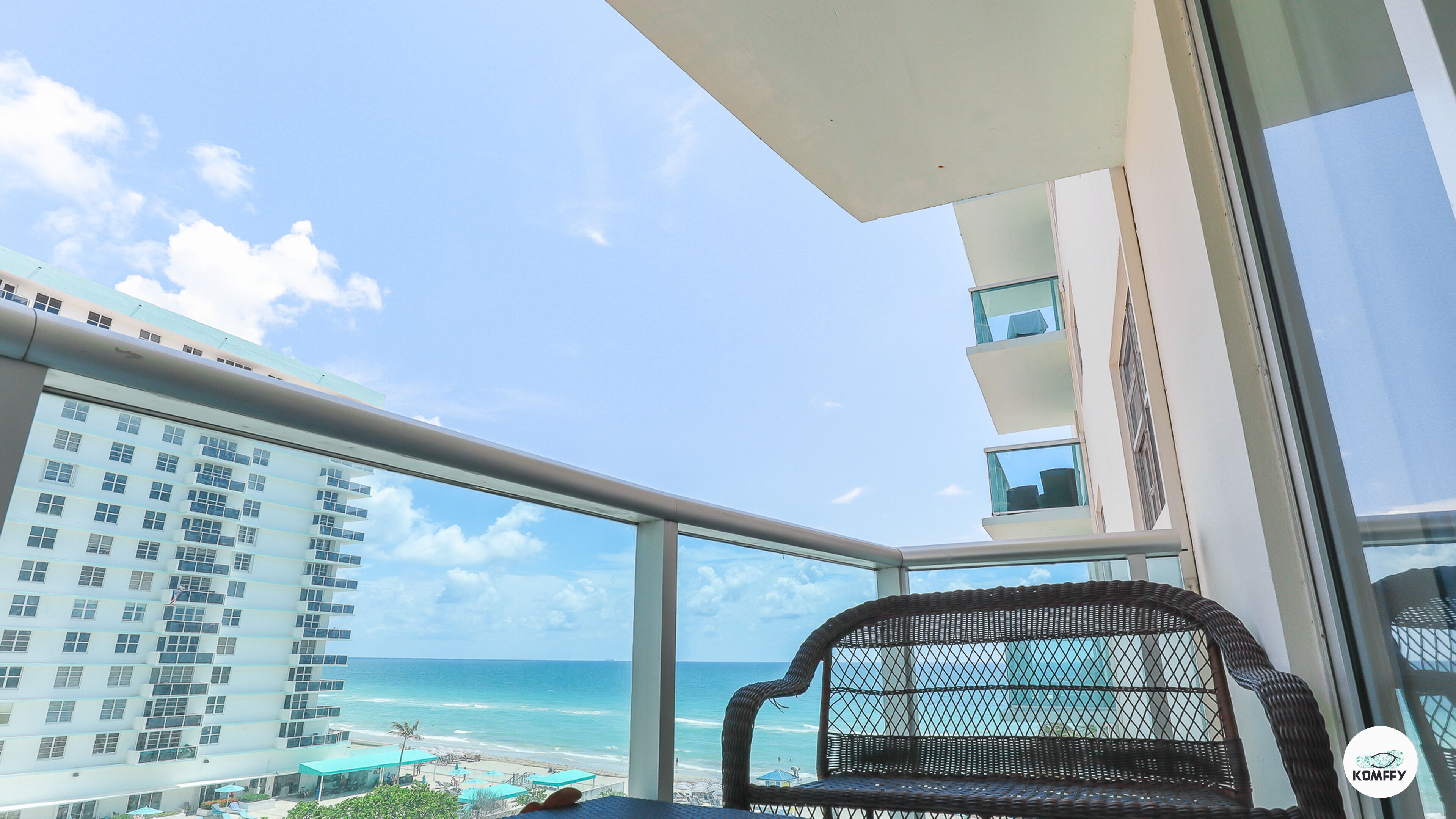 A balcony with a wicker chair, a view of the beach, and a high-rise building in the background.