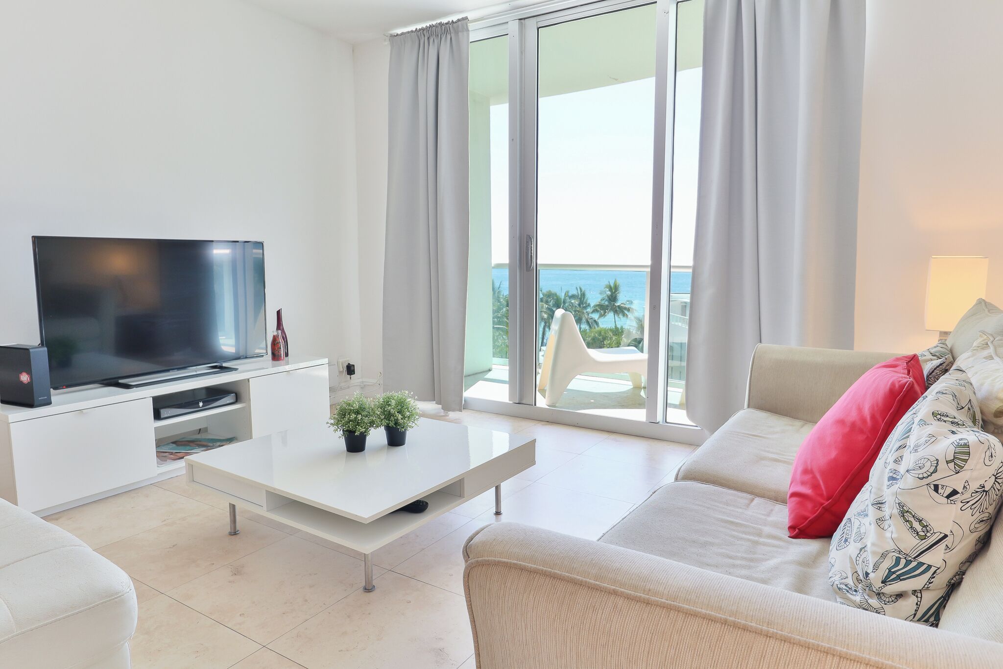 A modern living room with a flat-screen TV, a white coffee table with potted plants, a beige sofa with patterned cushions, and a balcony with a view of the ocean.