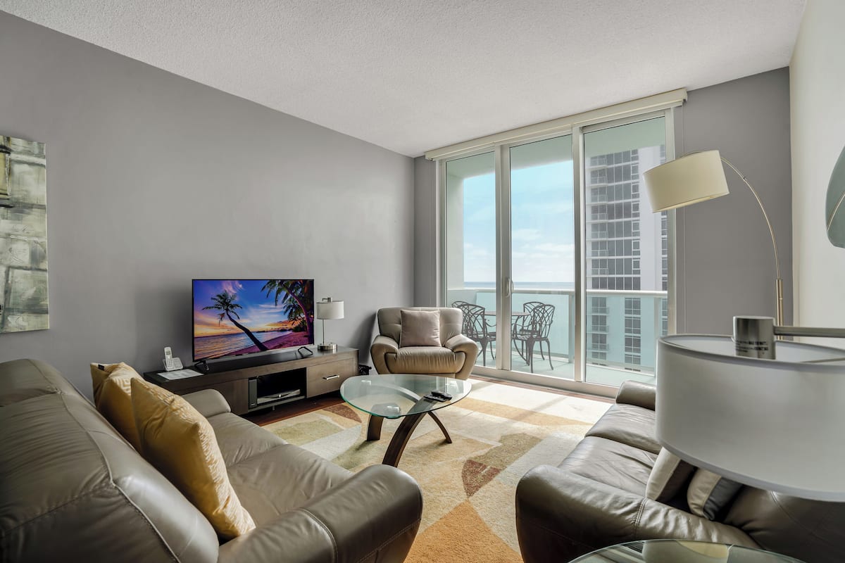 Una sala de estar moderna con un sofá, un sillón y una mesa de centro de vidrio. Un televisor de pantalla plana está sobre un mueble bajo, y hay un balcón con vistas.
