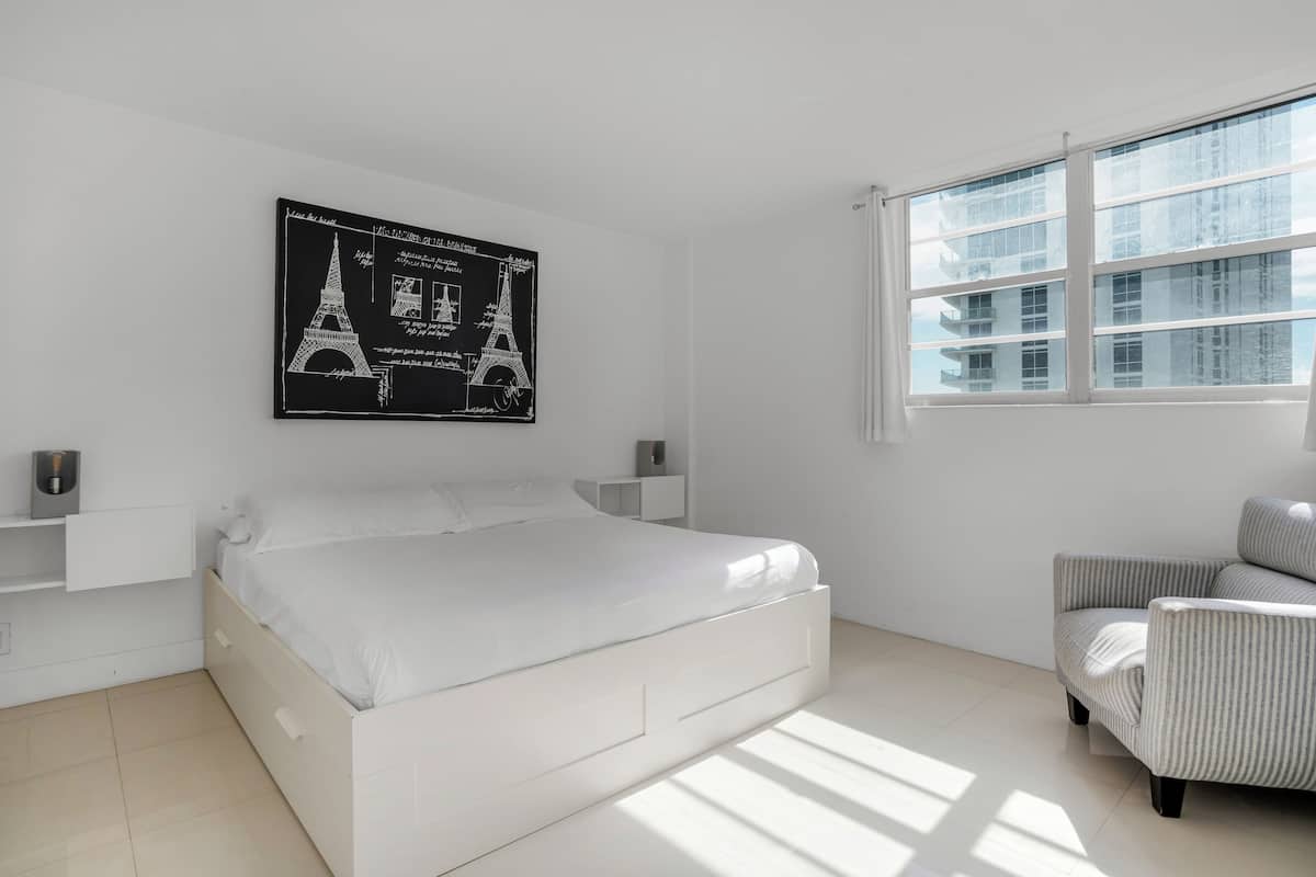 Un dormitorio moderno con una cama blanca, una mesita de noche, una silla, un ventanal con vistas a la ciudad y un póster blanco y negro de la Torre Eiffel en la pared.