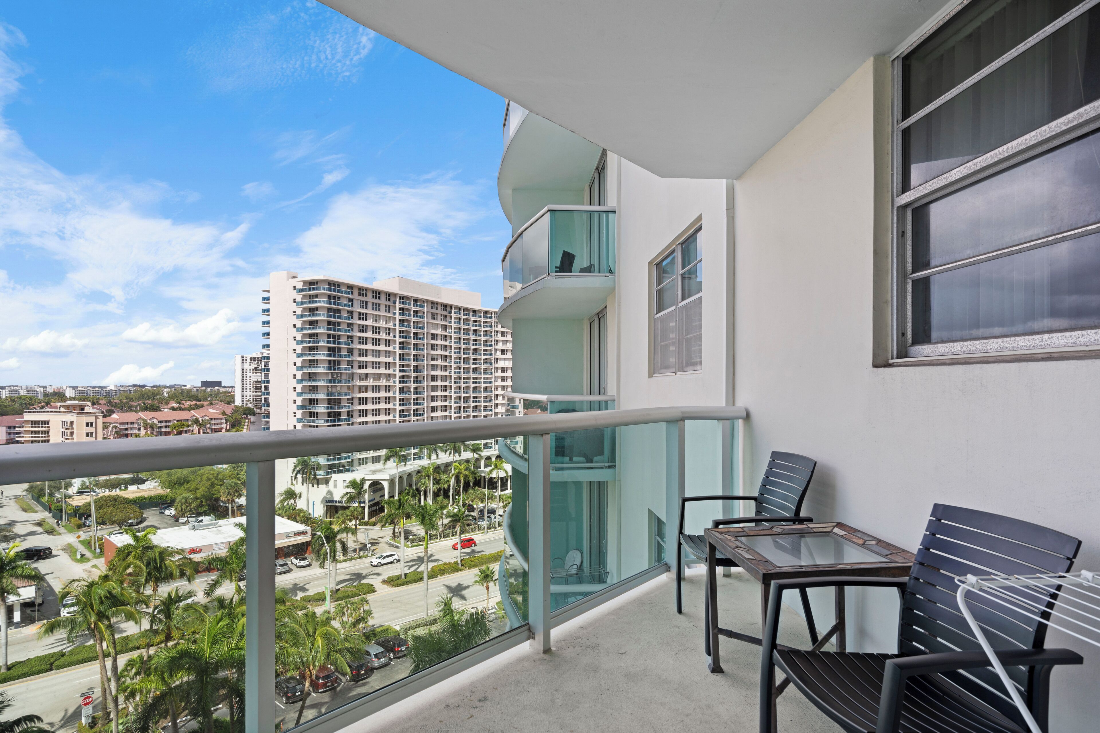 A balcony with a glass railing, a table with chairs, and a view of a cityscape with buildings and palm trees.