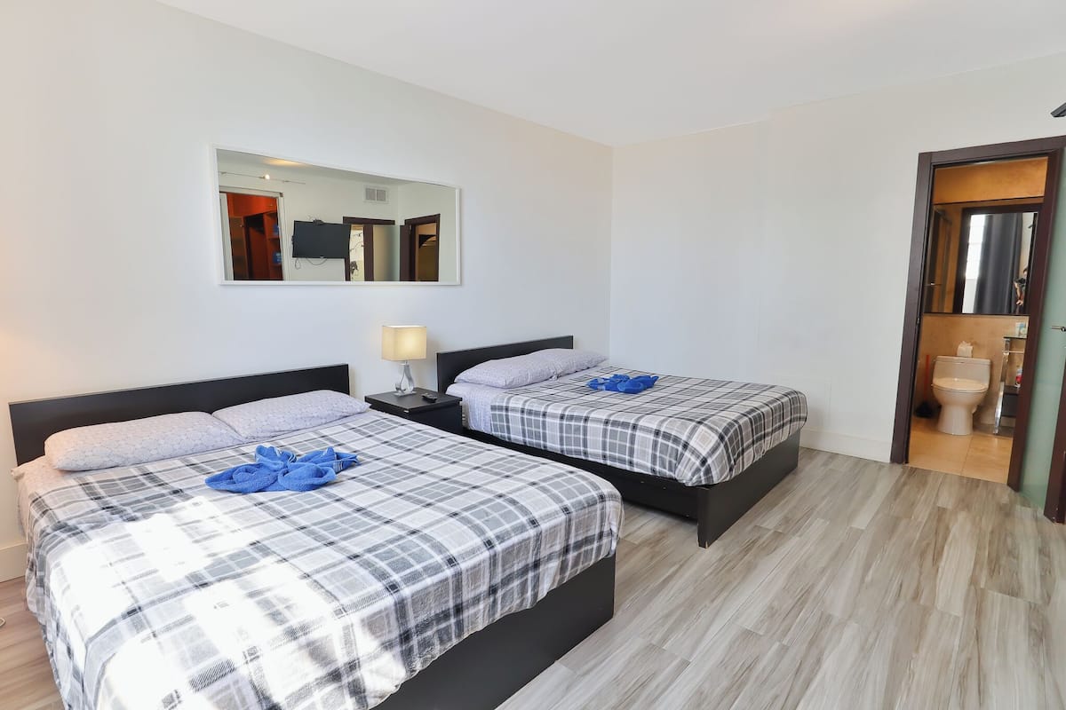 Habitación de hotel con dos camas, una mesita de noche, un espejo y un baño visible a través de una puerta abierta.