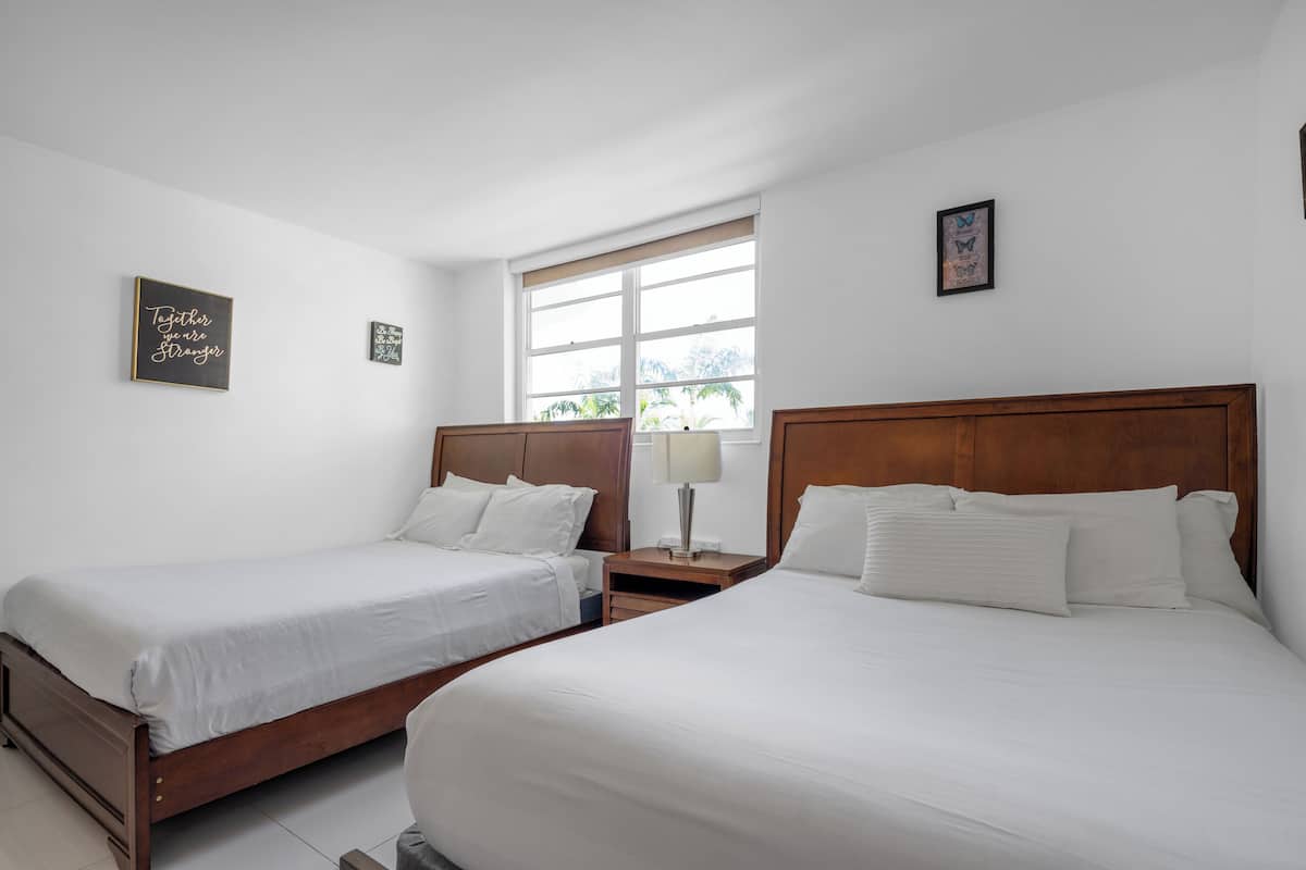 Una habitación de hotel con dos camas individuales, una mesita de noche con lámpara y una ventana con vistas a palmeras.