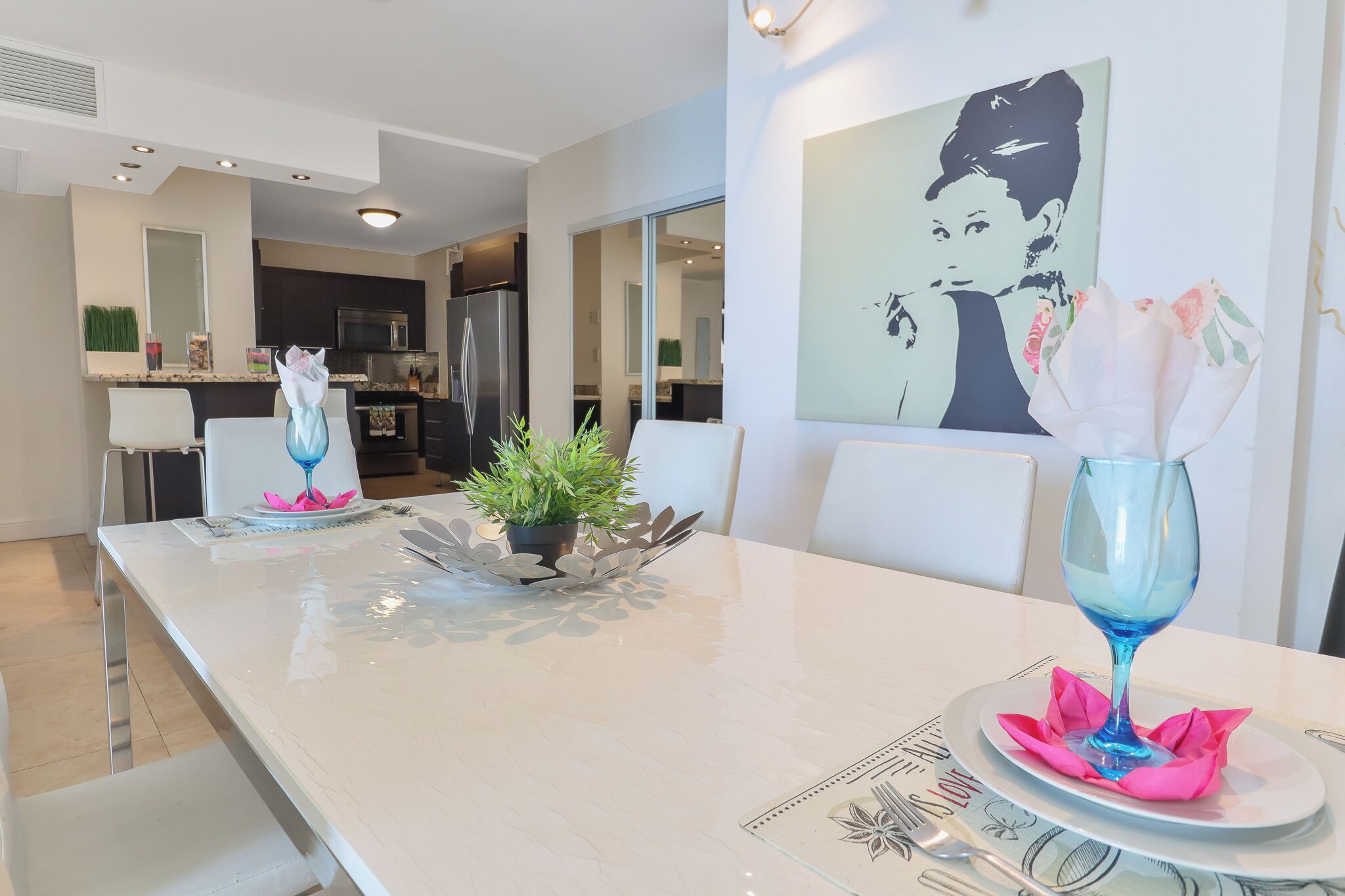 Un comedor moderno con una mesa blanca dispuesta para dos personas, destacando un vaso azul y una disposición de servilletas rosas.