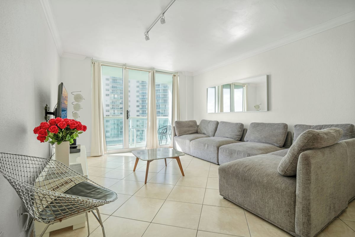 Una sala de estar moderna con un sofá gris, una silla de metal, una mesa de centro de vidrio y un jarrón con flores rojas.
