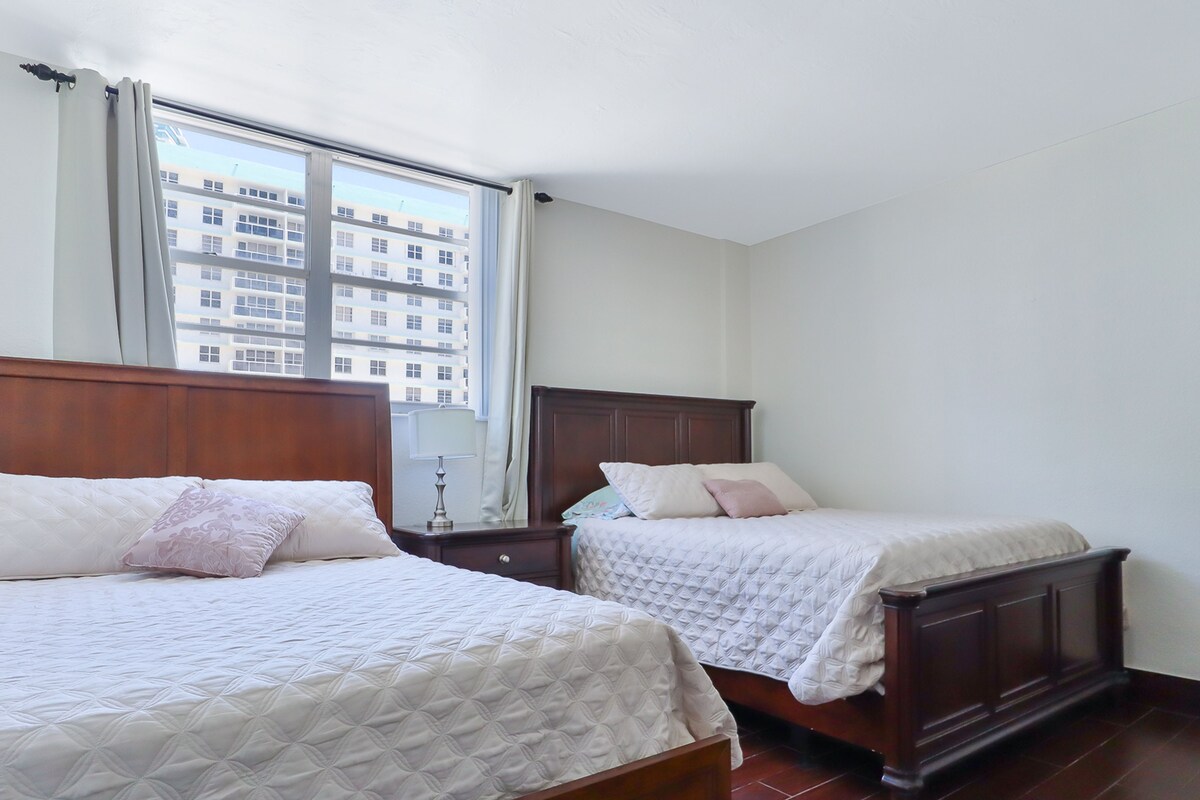 Habitación de hotel con dos camas, un ventanal grande y vistas a un edificio.