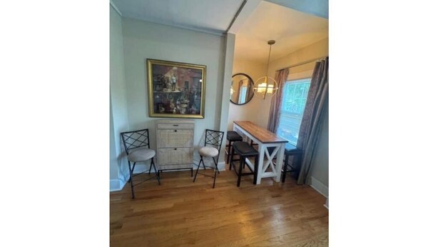 A room with a wooden floor, a dining table with chairs, a small cabinet, a framed picture on the wall, and a hanging light fixture.