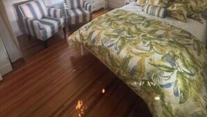 A bedroom with a bed, two chairs, a nightstand, a ceiling fan, and windows with curtains.
