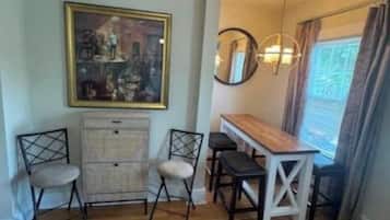 A room with a wooden floor, a dining table with chairs, a small cabinet, a framed picture on the wall, and a hanging light fixture.