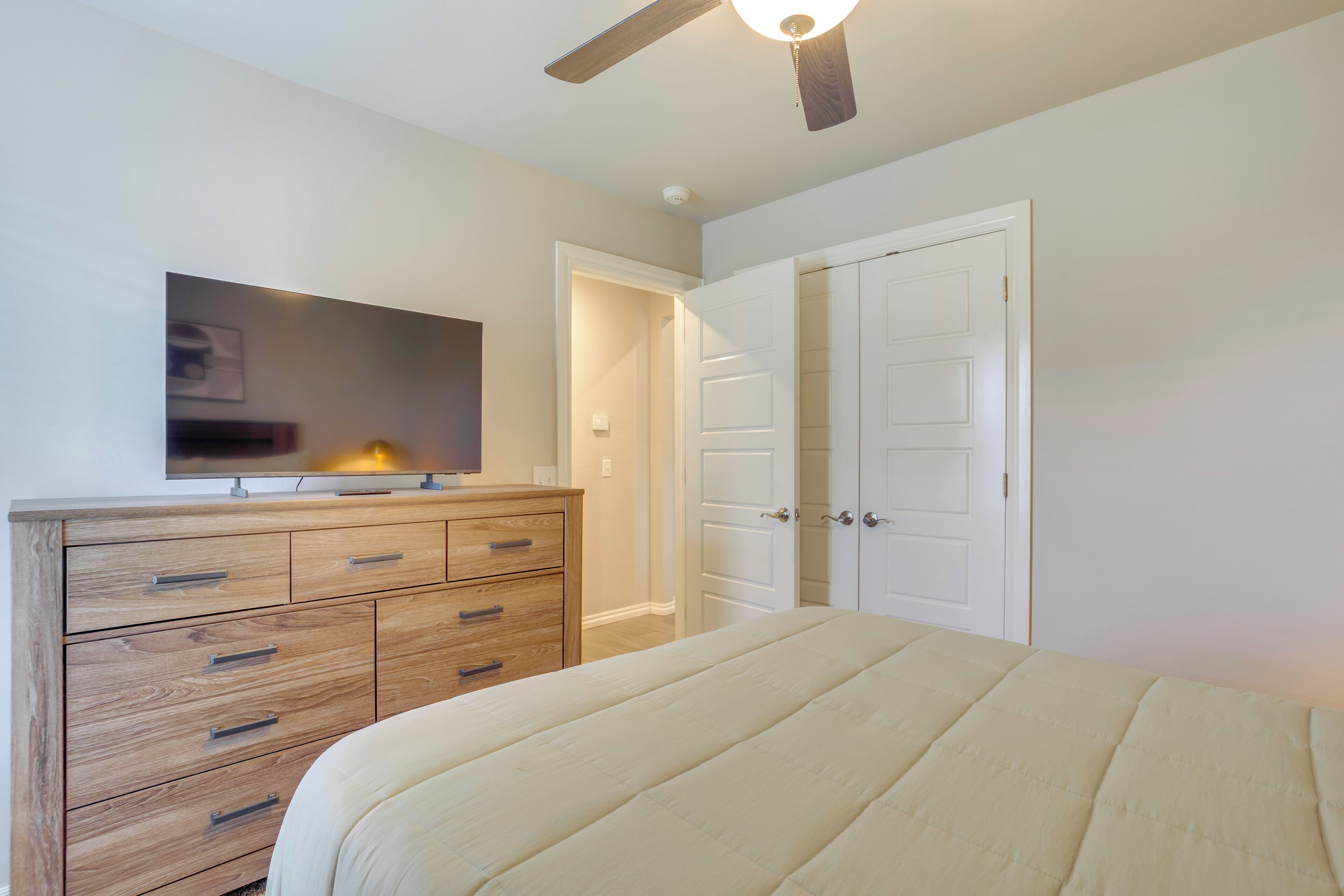 Une chambre à coucher comprenant un lit, une commode en bois, un ventilateur de plafond et un téléviseur fixé au mur.