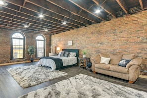Un dormitorio con pared de ladrillo, cama con cabecera verde, sofá color beige y dos ventanas grandes.