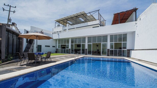 A modern outdoor pool area with a clear blue pool, a covered seating area with chairs and an umbrella, and a white building with large windows and a staircase. - House with Terrace & Pool in Coyula (Tonala)