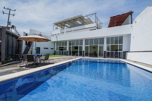 A modern outdoor pool area with a clear blue pool, a covered seating area with chairs and an umbrella, and a white building with large windows and a staircase. - House with Terrace & Pool in Coyula (Tonala)