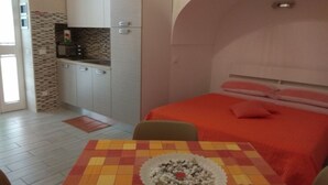 A compact room with a kitchenette, a dining area with a checkered tablecloth, and a bed with a red blanket.