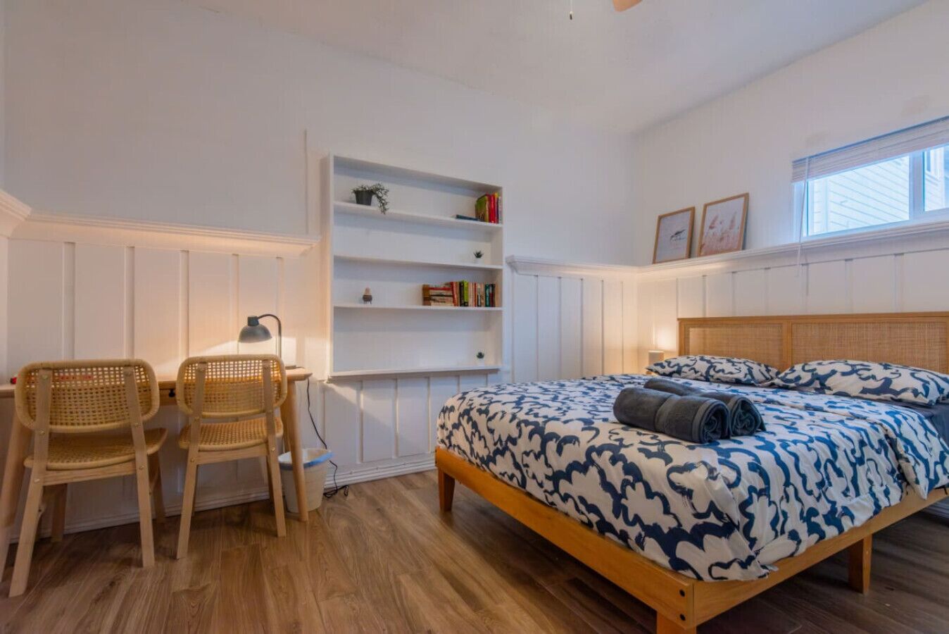 A bedroom with a wooden bed, two wicker chairs, a desk with a lamp, and built-in shelving with books and decorative items.