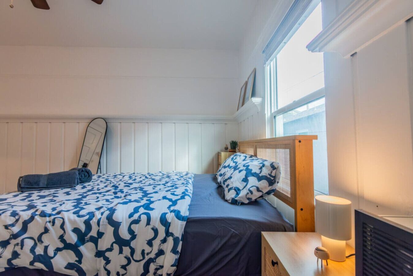 Ein Schlafzimmer mit einem Bett, einem Nachttisch mit Lampe, einem Fenster und einem Spiegel an der Wand.