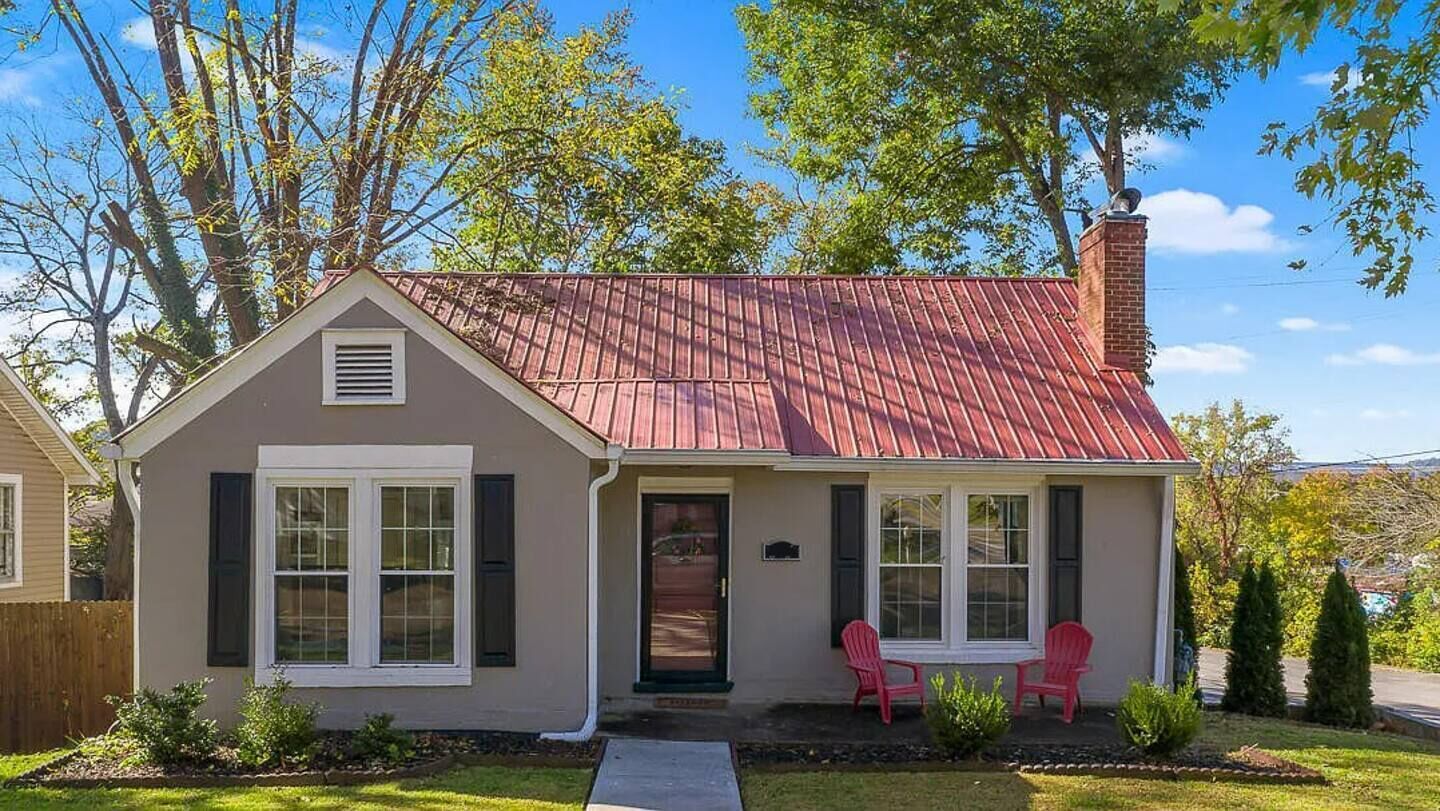 Uma casa térrea com telhado vermelho, varanda frontal com duas cadeiras vermelhas e um pequeno jardim.