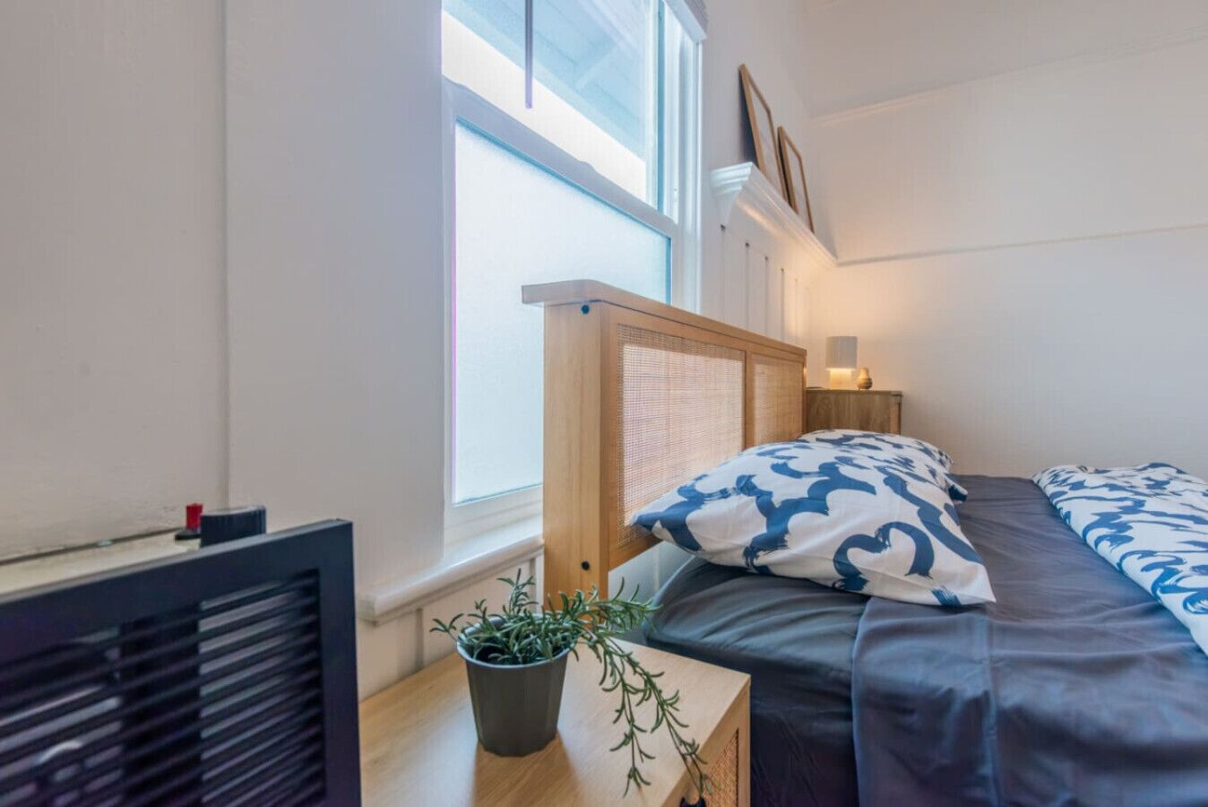 Et soveværelse med en seng, et sengebord med en plante, en radiator og et vindue med persienner.