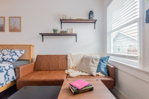 A living room with a brown leather sofa, a wooden coffee table with books, a window with blinds, and two wall-mounted shelves with decorative items. - Broadway 18 - Spacious Private Studio (Redondo Beach)