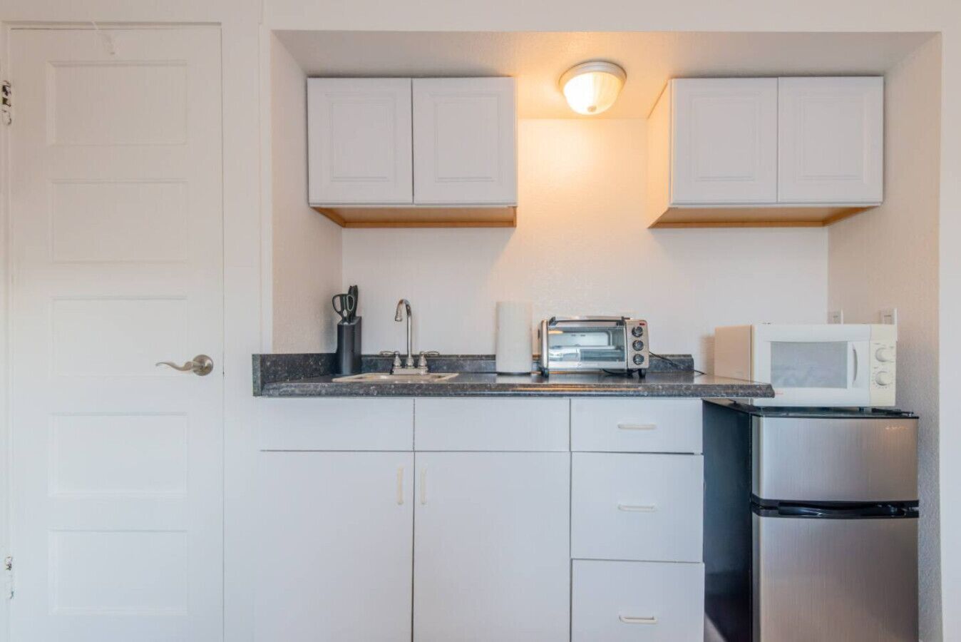 Een kleine keuken met witte kastjes, een roestvrijstalen koelkast, een broodrooster en een magnetron.