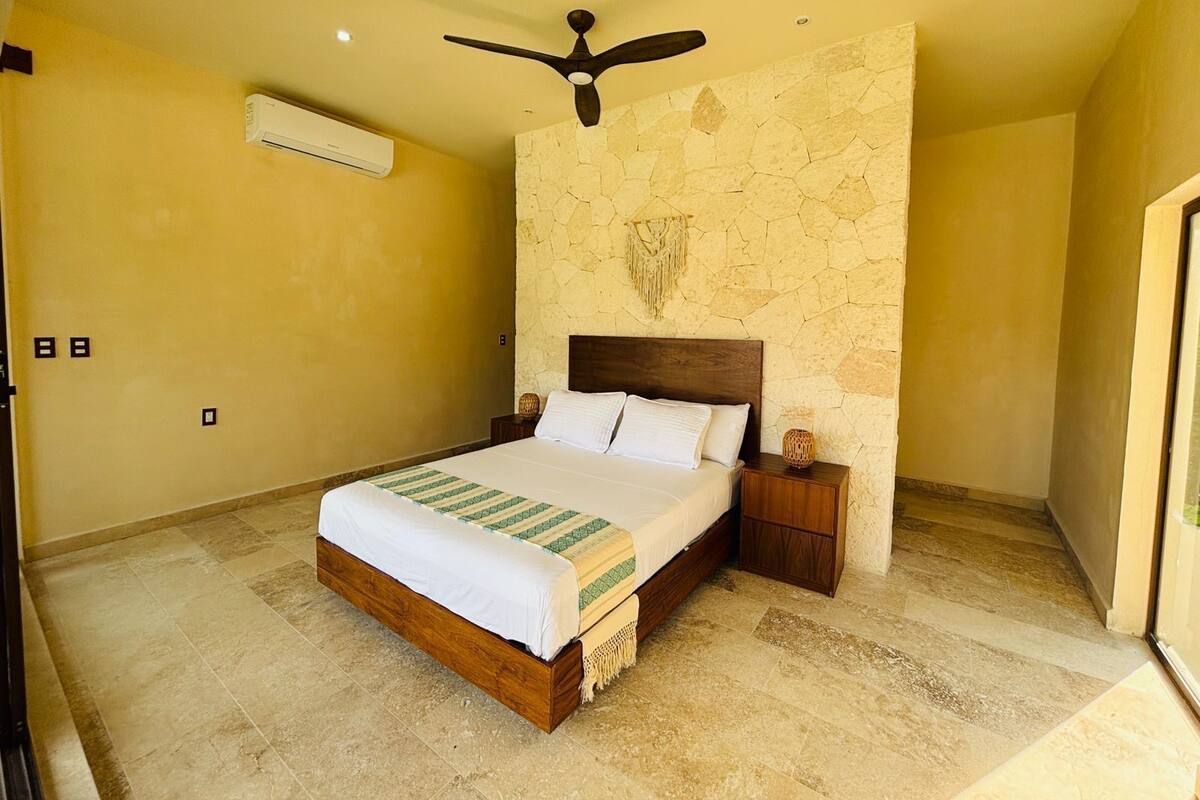 A bedroom with a stone wall, a wooden bed frame, a ceiling fan, and a nightstand with a lamp.